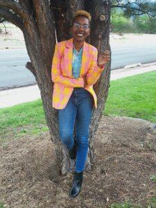 Miss Cadabra, smiling, leaning against a tree holding a joint in her hand