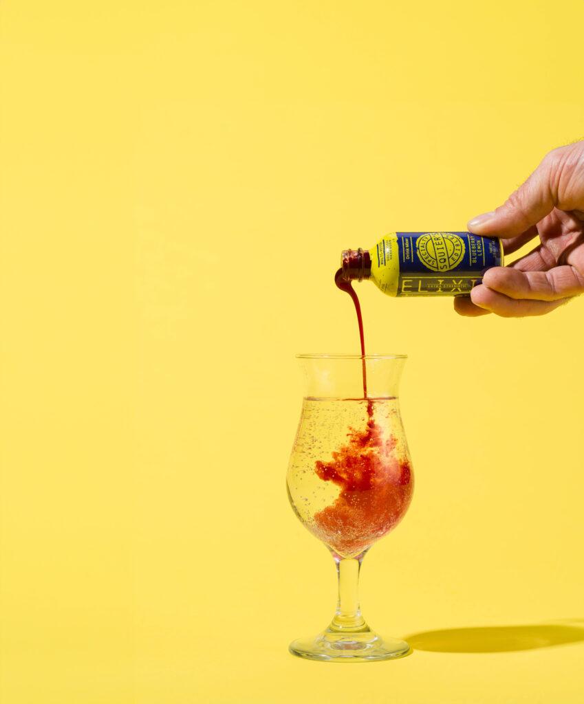 image of a bottle of Squier's Specialty Elixirs being poured in to a glass of clear liquid. 